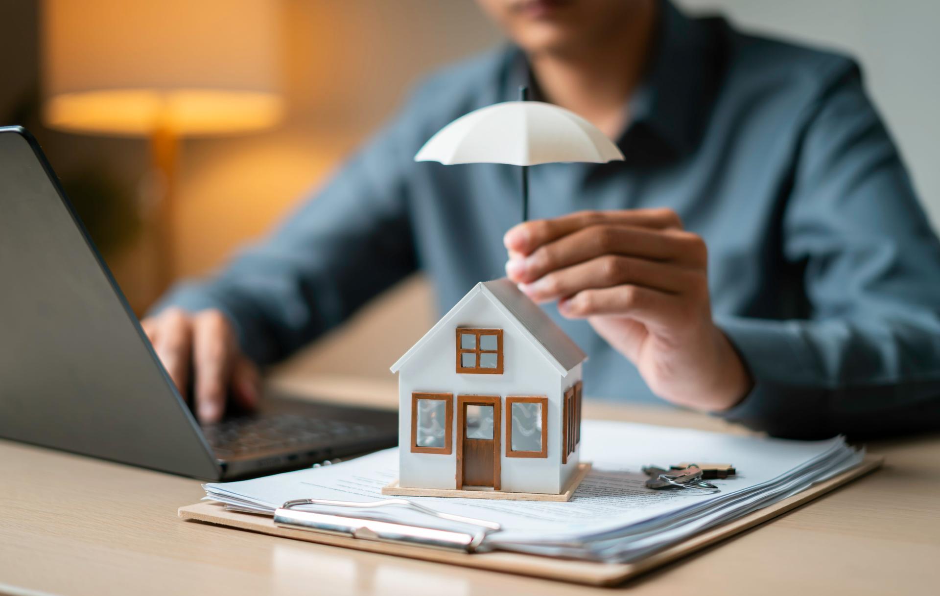 Close-up of a tiny house shielded by an umbrella atop loan documents next to keys and a laptop, representing property coverage.